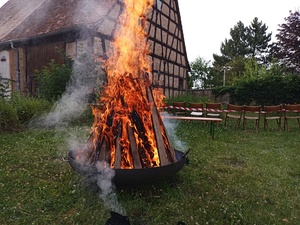 Im Vordergrund brennt ein Feuer in einer großen Feuerschale. Im Hintergrund ist eine schöne mit Fachwerk zu sehen.