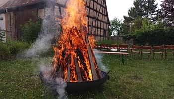 Im Vordergrund brennt ein Feuer in einer großen Feuerschale. Im Hintergrund ist eine schöne mit Fachwerk zu sehen.