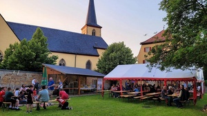 Auf einer grünen Wiese sind Bänke und ein kleines weißes Zelt aufgestellt. Menschen sitzen und stehen im Garten. Im Hintergrund ist eine Mauer und eine Kirche.