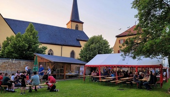 Auf einer grünen Wiese sind Bänke und ein kleines weißes Zelt aufgestellt. Menschen sitzen und stehen im Garten. Im Hintergrund ist eine Mauer und eine Kirche.