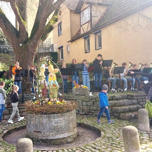 Um einen runden österlich geschmückten Steinbrunnen suchen Kinder etwas. Im Hintergrund spielt eine Musikgruppe.