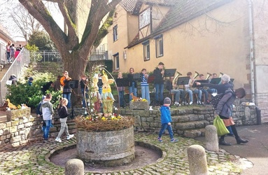 Um einen runden österlich geschmückten Steinbrunnen suchen Kinder etwas. Im Hintergrund spielt eine Musikgruppe.