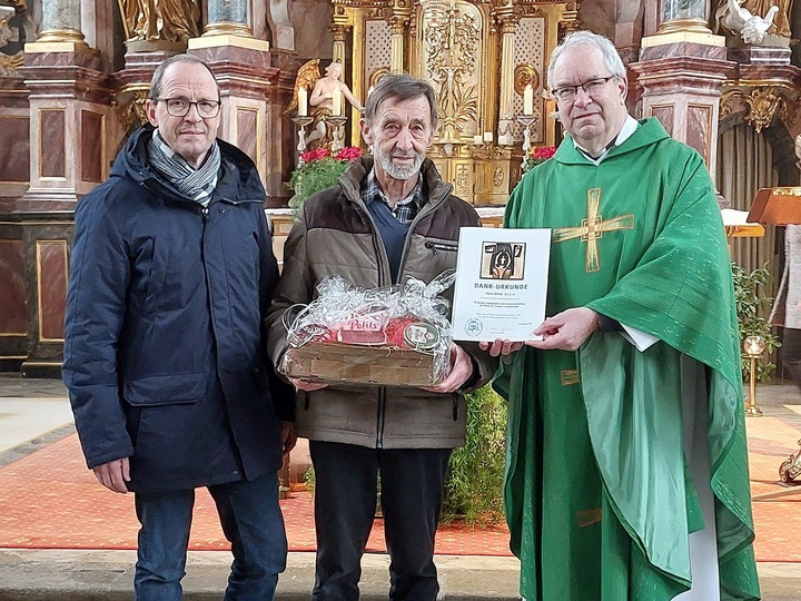 Drei Männer blicken in die Kamera. In der Mitte hält Alfred Stark den Präsentkorb in der Hand. Der Pfarrer zeigt die Dankurkunde.