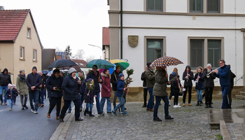 Mit einer Prozession ziehen Menschen in die Kirche ein. Es regnet, einige Regenschirme sind aufgespannt. Rechts steht eine Musikkapelle.