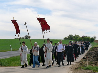 Ein Gruppe von Menschen, darunter Männer in weißen Gewändern und Frauen in dunklen Kleidern, geht auf einem Feldweg entlang einer Straße. Einige tragen rote Fahnen mit goldenen Kreuzen. Die Landschaft ist grün und offen, der Himmel blau mit Wolken.