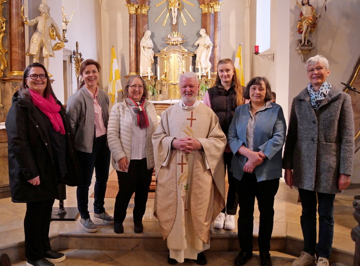 Das Bild zeigt eine Gruppe von sechs Personen, die in einer Kirche stehen. Im Zentrum befindet sich ein Geistlicher in liturgischer Kleidung, umgeben von fünf Frauen in Winterjacken. Im Hintergrund ist ein verzierter Altar mit Statuen und Kerzen zu sehen.
