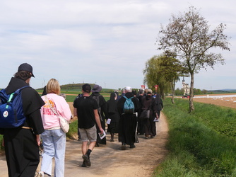 Das Bild zeigt eine Gruppe von Menschen, die auf einem Feldweg gehen. Einige tragen schwarze Kutten und andere legerere Kleidung. Im Hintergrund sind Felder und einige Bäume zu sehen, sowie eine entfernte Kirche. Der Himmel ist klar.