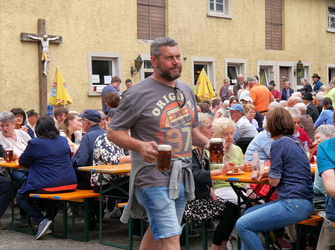 Auf dem Bild sieht man eine gesellige Veranstaltung im Freien mit vielen Menschen an Tischen, die Bier trinken. Ein bärtiger Mann in kurzen Hosen steht im Vordergrund. Häuser und Fahnen sind im Hintergrund.