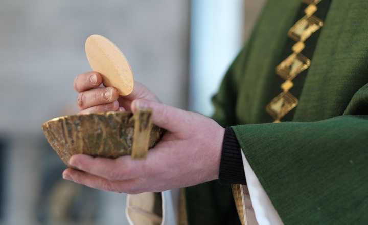 Mit der linken Hand hält ein Priester eine Hostienschale. Mit der rechten Hand hält er darüber eine große Hostie.