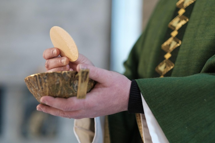 Mit der linken Hand hält ein Priester eine Hostienschale. Mit der rechten Hand hält er darüber eine große Hostie.