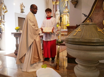 Das Bild zeigt einen Altarraum in einer Kirche. Ein Priester im weißen Gewand hält ein Buch, während ein junger Assistent in weißem Gewand mit rotem Akzent daneben steht. Im Hintergrund sind ein verzierter Altar und ein Taufbecken zu sehen.