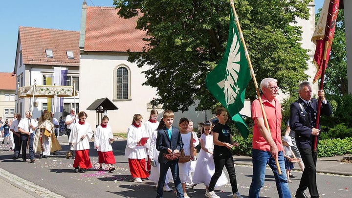 Fronleichnamsprozession mit Fahnenträgern voran und dem Himmel und dem Allerheiligsten im Hintergrund.