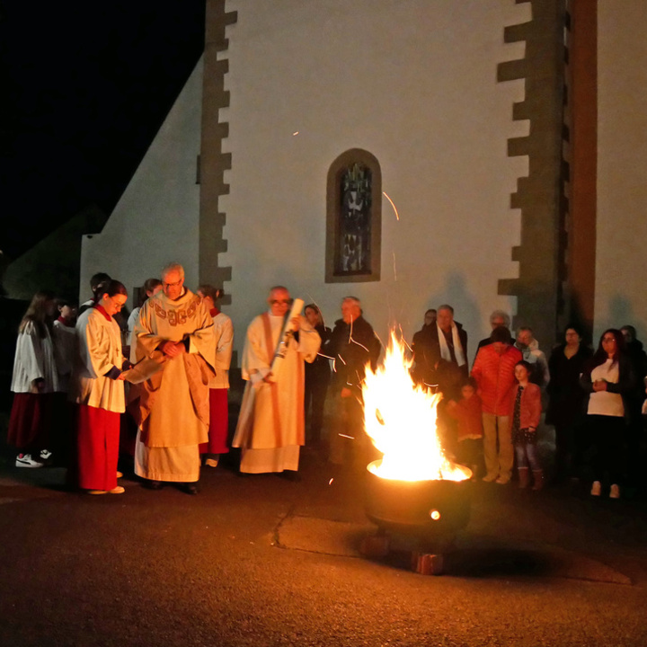 Ein Osterfeuer brennt auf dem Kirchplatz in Dipbach. Die Menschen, die dahinter stehen, sind vom Feuerschein erhellt.