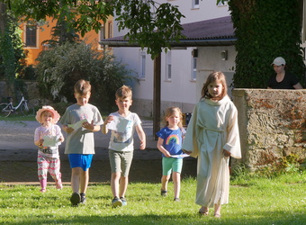 Auf dem Bild laufen fünf Kinder und eine erwachsene Frau in leichter Kleidung auf einem Rasen vor einem Haus mit Papieren in den Händen. Bäume und Fahrräder sind sichtbar.