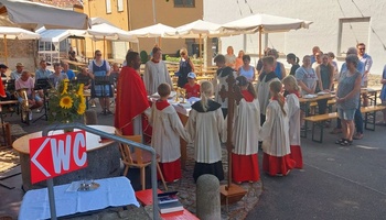 Das Bild zeigt einen Priester und Ministranten, die sich bei einem Gottesdienst auf dem Dorfplatz um den Altar versammeln.