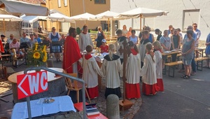 Das Bild zeigt einen Priester und Ministranten, die sich bei einem Gottesdienst auf dem Dorfplatz um den Altar versammeln.