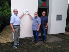 Drei Männer stehen an der Außenwand einer Kapelle. Zwei von ihnen halten ein rundes Schild hoch mit der Aufschrift 