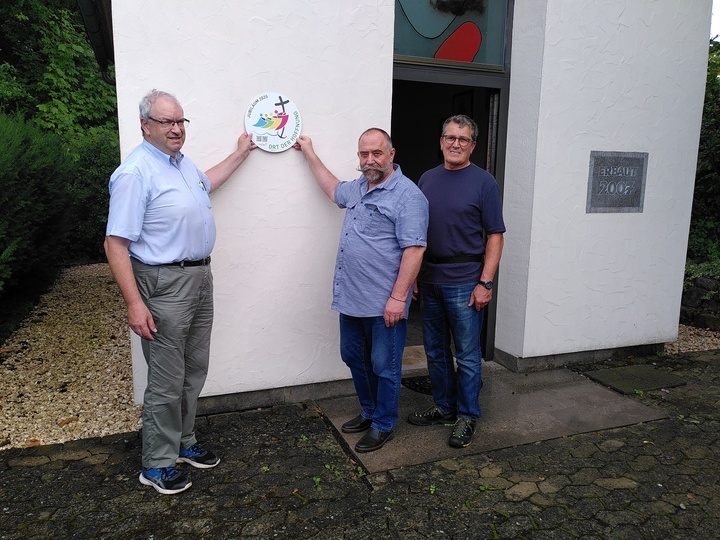 Drei Männer stehen an der Außenwand einer Kapelle. Zwei von ihnen halten ein rundes Schild hoch mit der Aufschrift "Ort der Hoffnung".