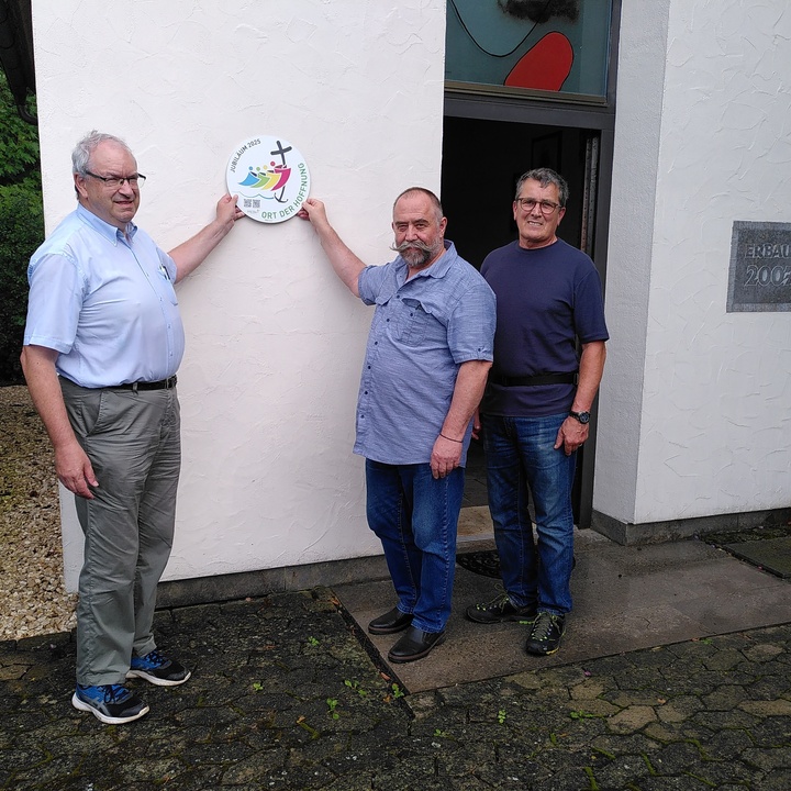 Drei Männer stehen an der Außenwand einer Kapelle. Zwei von ihnen halten ein rundes Schild hoch mit der Aufschrift "Ort der Hoffnung".