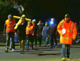 Das Bild zeigt eine Gruppe von Menschen, die nachts eine Prozession abhalten. Einige tragen Warnwesten und einer hält ein großes gelbes Kreuz. Die Szene ist dunkel und wird von Taschenlampen beleuchtet.