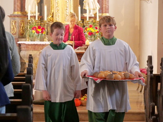 Das Bild zeigt zwei Ministranten im Mittelgang einer Kirche. Der rechte Junge hat ein Tablet in der Hand, auf dem ein Brot liegt.