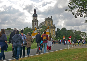 Im Mittelpunkt des Bildes ist die Wallfahrtskirche Fährbrück von außen. Links und rechts sieht man jeweils eine Prozession auf die Kirche zugehen.