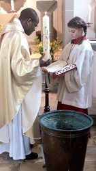 Auf dem Bild sind ein Pfarrer mit der Osterkerze und ein Ministrant mit dem Messbuch zu sehen. Vor ihnen steht ein Behälter mit Wasser.