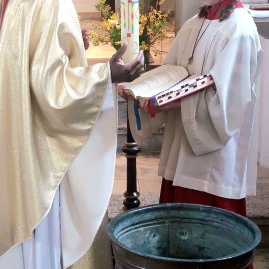 Auf dem Bild sind ein Pfarrer mit der Osterkerze und ein Ministrant mit dem Messbuch zu sehen. Vor ihnen steht ein Behälter mit Wasser.