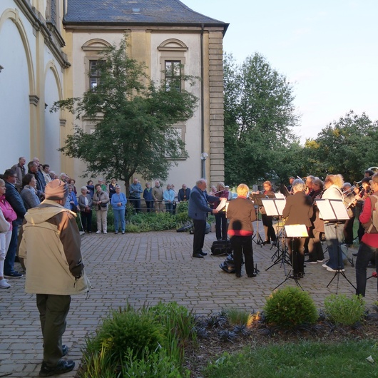 Am Seitenausgang der Wallfahrtskirche spielt rechts die Musikkapelle. Um sie herum stehen Menschen und hören zu.