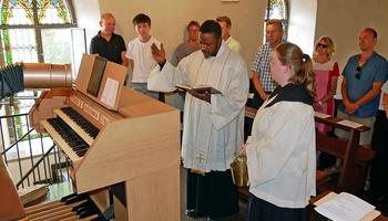 Ein Priester steht vor dem Spieltisch einer Orgel und spricht ein Segensgebet. Um ihn herum stehen weitere Menschen.