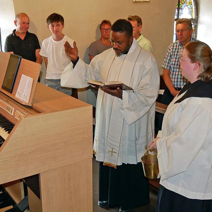 Ein Priester steht vor dem Spieltisch einer Orgel und spricht ein Segensgebet. Um ihn herum stehen weitere Menschen.