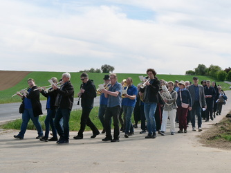 Das Bild zeigt eine Gruppe von Menschen, die auf einer Landstraße bei einer Wallfahrt. Einige von ihnen spielen Blasinstrumente wie Trompeten und Tubas. Die Umgebung ist ländlich mit grünen Feldern und einem blauen Himmel.
