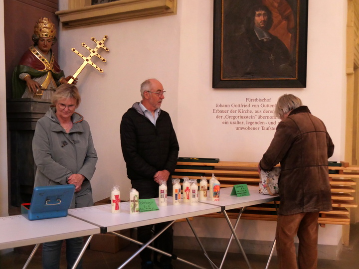 Hinter einem Verkaufstisch mit Osterkerzen stehen eine Frau und ein Mann. Davor schaut eine Frau die Kerzen an. Diese Frau sieht man nur von hinten.