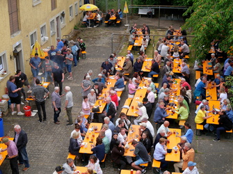 Auf dem Bild sieht man viele Menschen, die in einem Innenhof an gelben Tischen sitzen und essen. Es wirkt wie ein geselliges Fest oder eine öffentliche Veranstaltung.