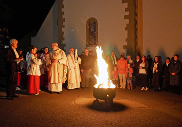Auf dem Kirchplatz brennt hell das Osterfeuer. Pfarrer Rügamer betet etwas und liest es aus einem Buch vor.