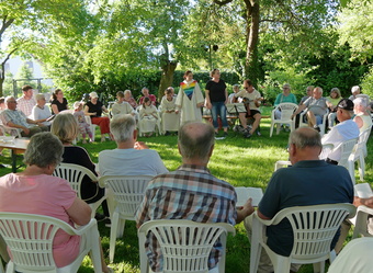 Eine Gruppe von Menschen sitzt im Freien auf weißen Stühlen unter Bäumen und nimmt an einer Zeremonie mit einer Person in liturgischer Kleidung teil.