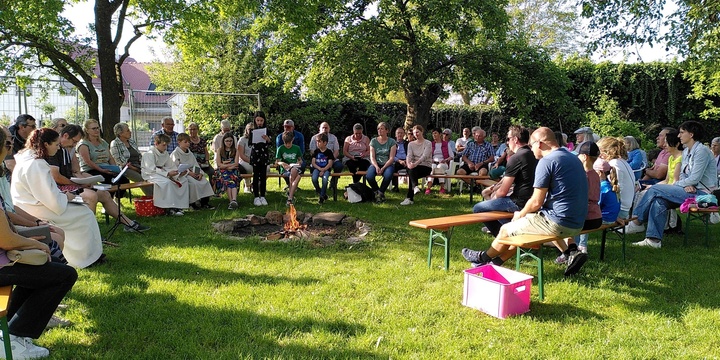 Im Grünen und im Kreisrund sitzen Menschen auf Bierbänken. Links sitzt Susanne Händel in einem weißen liturgischen Gewand.