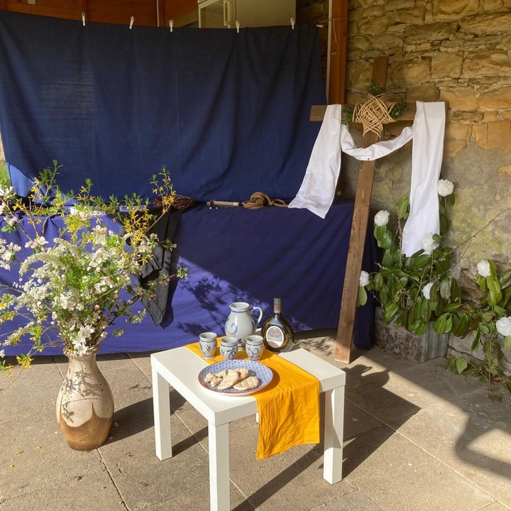 Im Vordergrund steht ein kleiner Tisch, auf dem ein Teller mit Brot und eine Weinflasche stehen. Links neben dem Tisch steht ein Blumenstrauß in einer Vase. Im Hintergrund ist ein großes blaues Tuch gespannt, vor dem ein Kreuz mit einem weißen Tuch steht