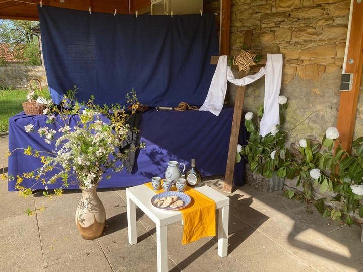 Im Vordergrund steht ein kleiner Tisch, auf dem ein Teller mit Brot und eine Weinflasche stehen. Links neben dem Tisch steht ein Blumenstrauß in einer Vase. Im Hintergrund ist ein großes blaues Tuch gespannt, vor dem ein Kreuz mit einem weißen Tuch steht