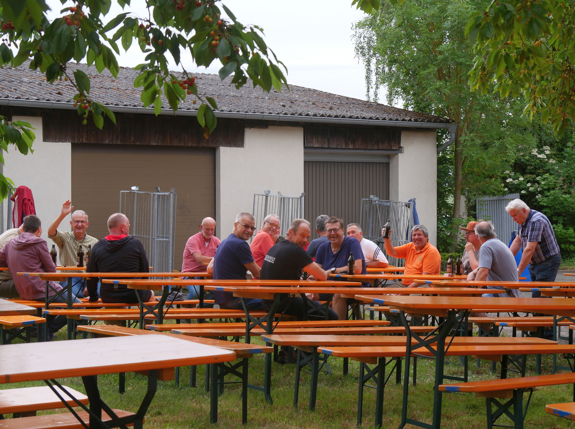 Auf dem Bild sitzen mehrere Männer an langen Holzbänken unter Bäumen vor einem Haus und unterhalten sich fröhlich bei einem geselligen Beisammensein.