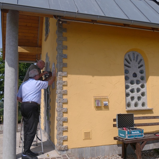 Die zwei Männer arbeiten gerade am Anbringen des Schildes. Die Kapelle ist ein Stückweit mit auf dem Bild.