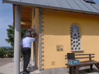 Die zwei Männer arbeiten gerade am Anbringen des Schildes. Die Kapelle ist ein Stückweit mit auf dem Bild.