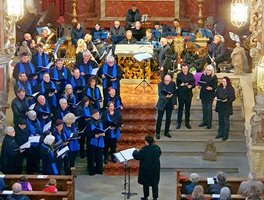 Das Bild zeigt einen Chor und ein Orchester in einer Kirche während einer Aufführung. Die Chormitglieder tragen schwarze Kleidung mit blauen Schals und halten Notenblätter. Im Hintergrund sitzen Musiker mit verschiedenen Instrumenten. Ein Dirigent steht vorne und leitet die Aufführung.
