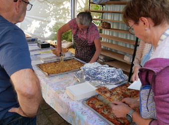 Das Bild zeigt einen Verkaufstisch. Rechts schneiden zwei Frauen Pizza und Zwiebelplotz auf. Links wartet ein Mann auf seine Bestellung.