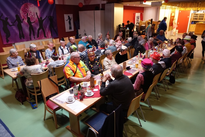 40 Menschen sitzen an zwei langen Tischreihen. Die Senioren sind verkleidet mit bunten Hüten und Kostümen, sie unterhalten sich und auf den Tischen stehen Getränke.