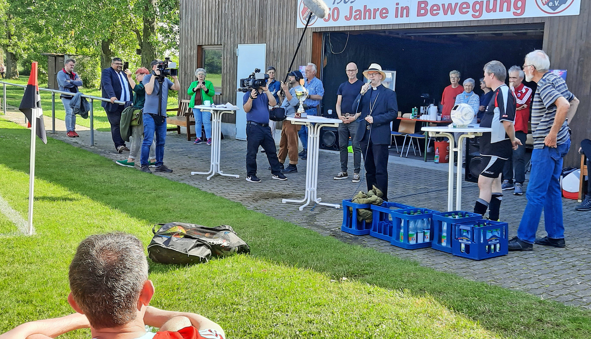 Neben einem Fußballfeld steht eine Gruppe Menschen um drei weiße Tische herum. In der Mitte ist Bischof Franz Jung. Er hat ein Mikrofon in der Hand.