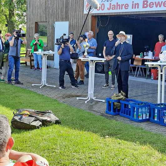 Neben einem Fußballfeld steht eine Gruppe Menschen um drei weiße Tische herum. In der Mitte ist Bischof Franz Jung. Er hat ein Mikrofon in der Hand.