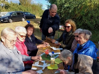 An einem Biertisch im Freien sitzen Menschen beieinander. Hinten am Tisch steht der Pastoralreferent Meier.