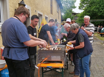 Auf dem Bild grillen mehrere Personen Würstchen an einem Gemeinschaftsgrillstand. Es herrscht eine gesellige Atmosphäre mit Menschen unterschiedlichen Alters im Freien.