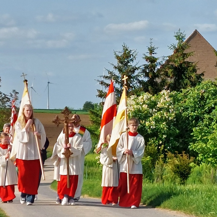 Ministrant:innen gehen in der Gruppe mit Prozessionsfahnen und Kreuz auf den Gottesdienstort zu.